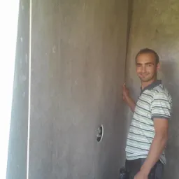 Mężczyzna w koszulce w paski uśmiecha się, opierając rękę na dużej, szarej płycie ściennej z otworem na instalację w trakcie montażu w łazience.