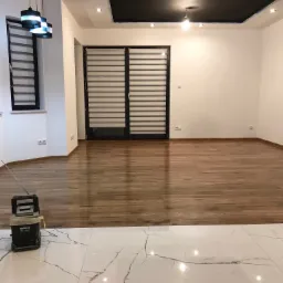 Wykończone wnętrze: połączenie drewnopodobnej podłogi i białych płytek imitujących marmur, widoczne lampy wiszące i okno z żaluzjami.