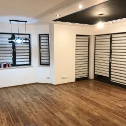 Puste, jasne pomieszczenie z drewnianą podłogą, trzema oknami z żaluzjami poziomymi i czarnym, podwieszanym sufitem z oświetleniem punktowym.