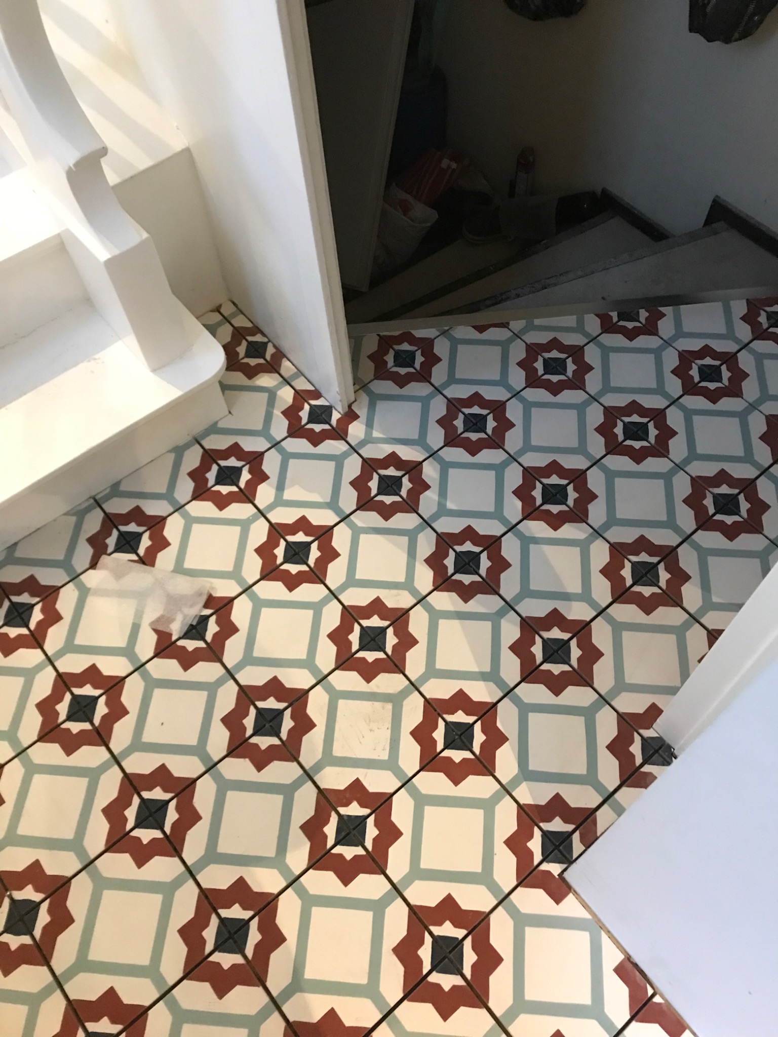 Podłoga w przedpokoju wyłożona wzorzystymi, kolorowymi płytkami ceramicznymi, z widocznym fragmentem białych schodów i wejściem do ciemnego pomieszczenia.