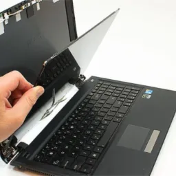 Otwarty laptop z odłączanym ekranem, widoczne wewnętrzne elementy i ręka osoby odczepiającej wyświetlacz od obudowy, białe tło.