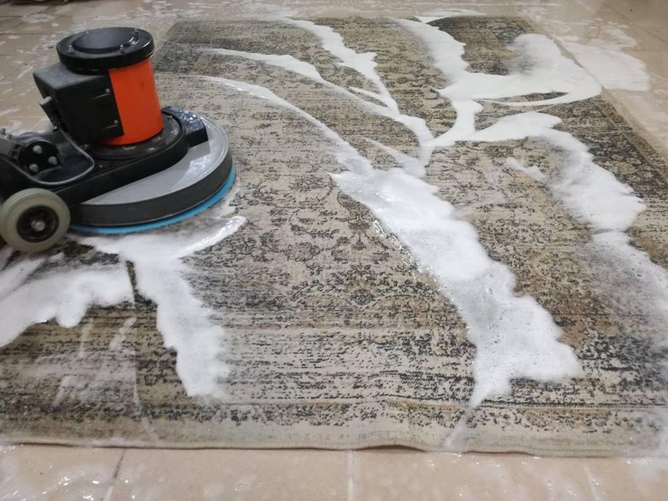 Szorowarka rotacyjna z pomarańczowym zbiornikiem czyści wzorzysty dywan, pozostawiając na nim grubą warstwę piany czyszczącej.