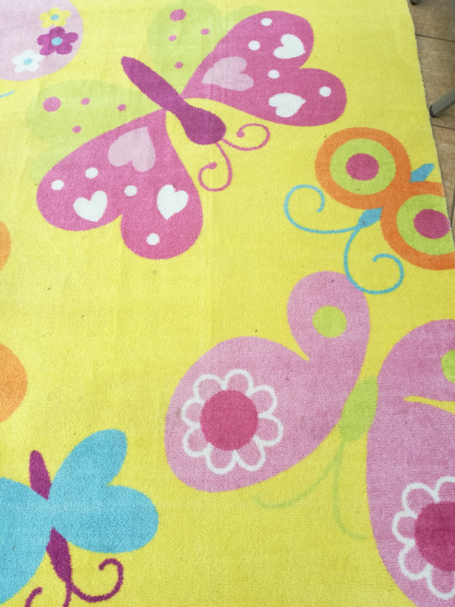 Żółty dywan z wzorem motyli i kwiatów w odcieniach różu, fioletu i niebieskiego, widoczne włosie dywanu.