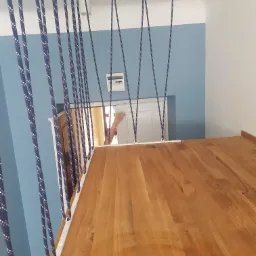 Antresola z drewnianym blatem, zabezpieczona sznurami w kolorze granatowym z białymi kropkami, widok z góry na jasną podłogę i błękitną ścianę.