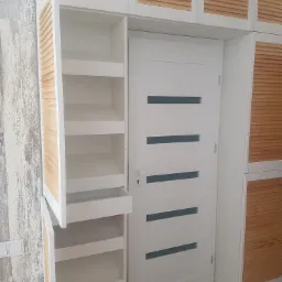 Biała szafa wnękowa z drewnianymi żaluzjowymi frontami, zintegrowana z białymi drzwiami wewnętrznymi z poziomymi przeszkleniami, widoczne półki w otwartej części szafy.