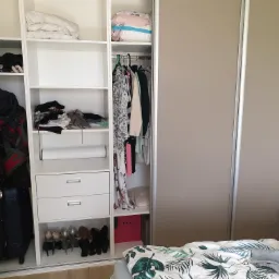 Wnętrze szafy wnękowej z przesuwnymi drzwiami w odcieniach beżu, widoczne półki z ubraniami, walizką, plecakiem i butami, białe szuflady i pudełko.