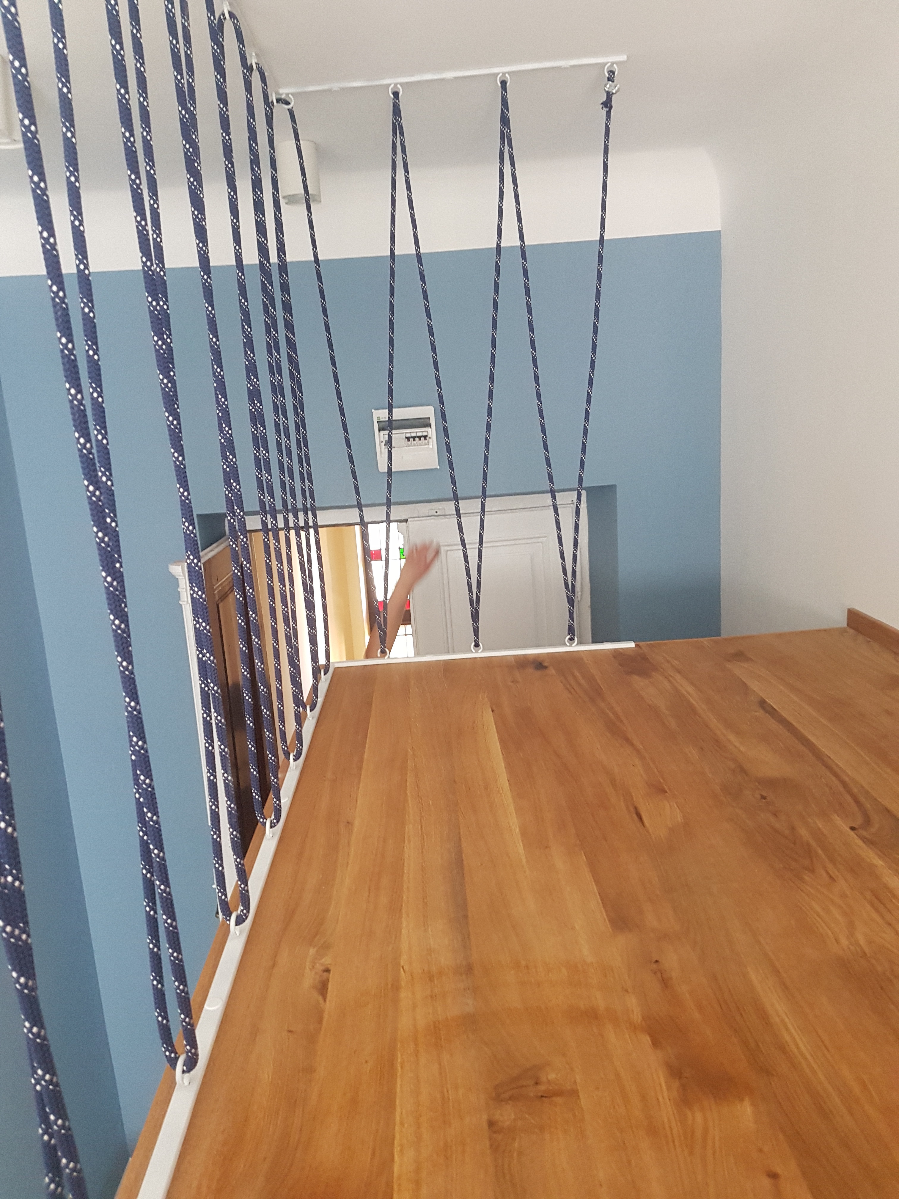 Antresola z drewnianym blatem, zabezpieczona sznurami w kolorze granatowym z białymi kropkami, widok z góry na jasną podłogę i błękitną ścianę.