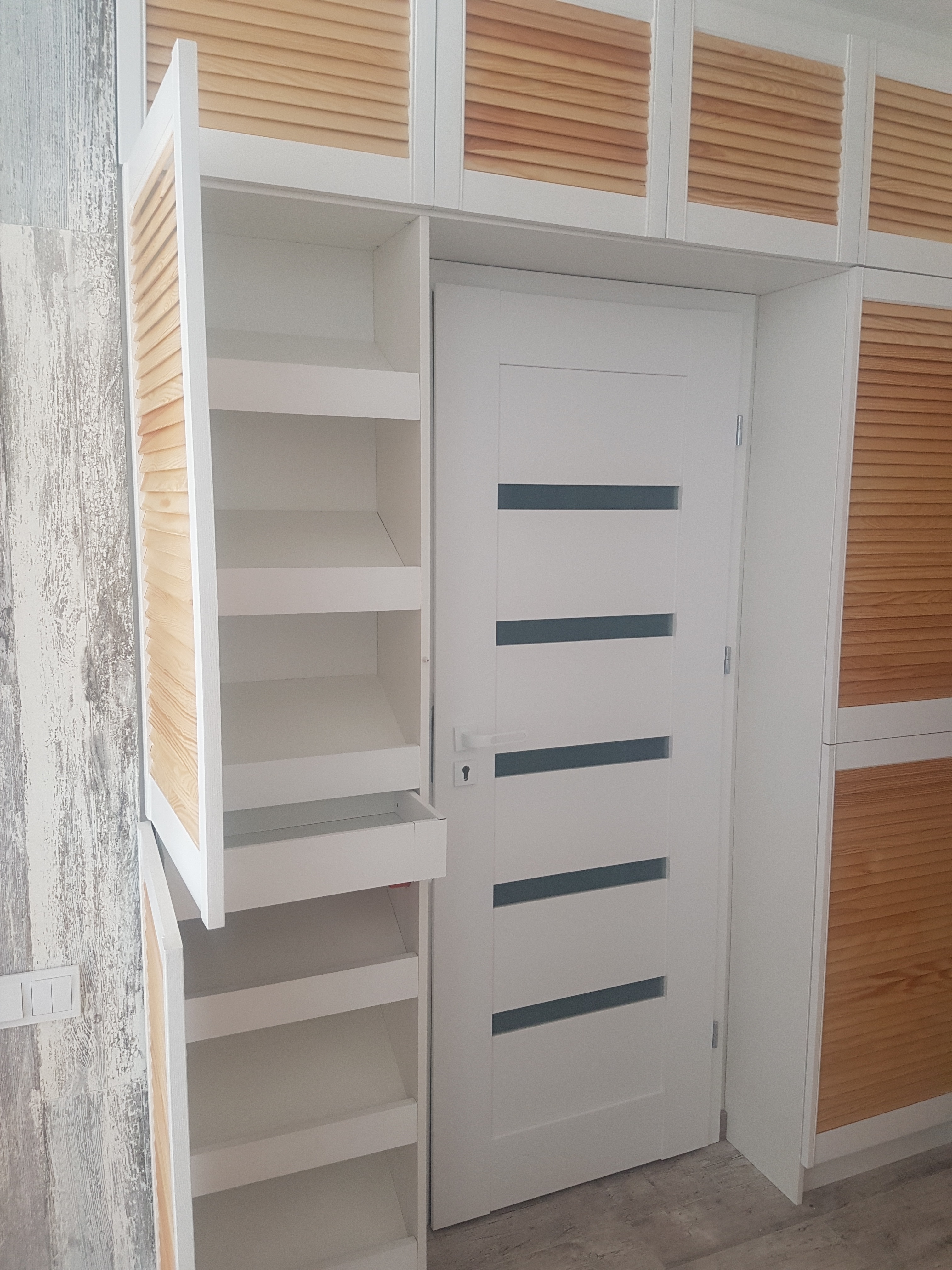 Biała szafa wnękowa z drewnianymi żaluzjowymi frontami, zintegrowana z białymi drzwiami wewnętrznymi z poziomymi przeszkleniami, widoczne półki w otwartej części szafy.