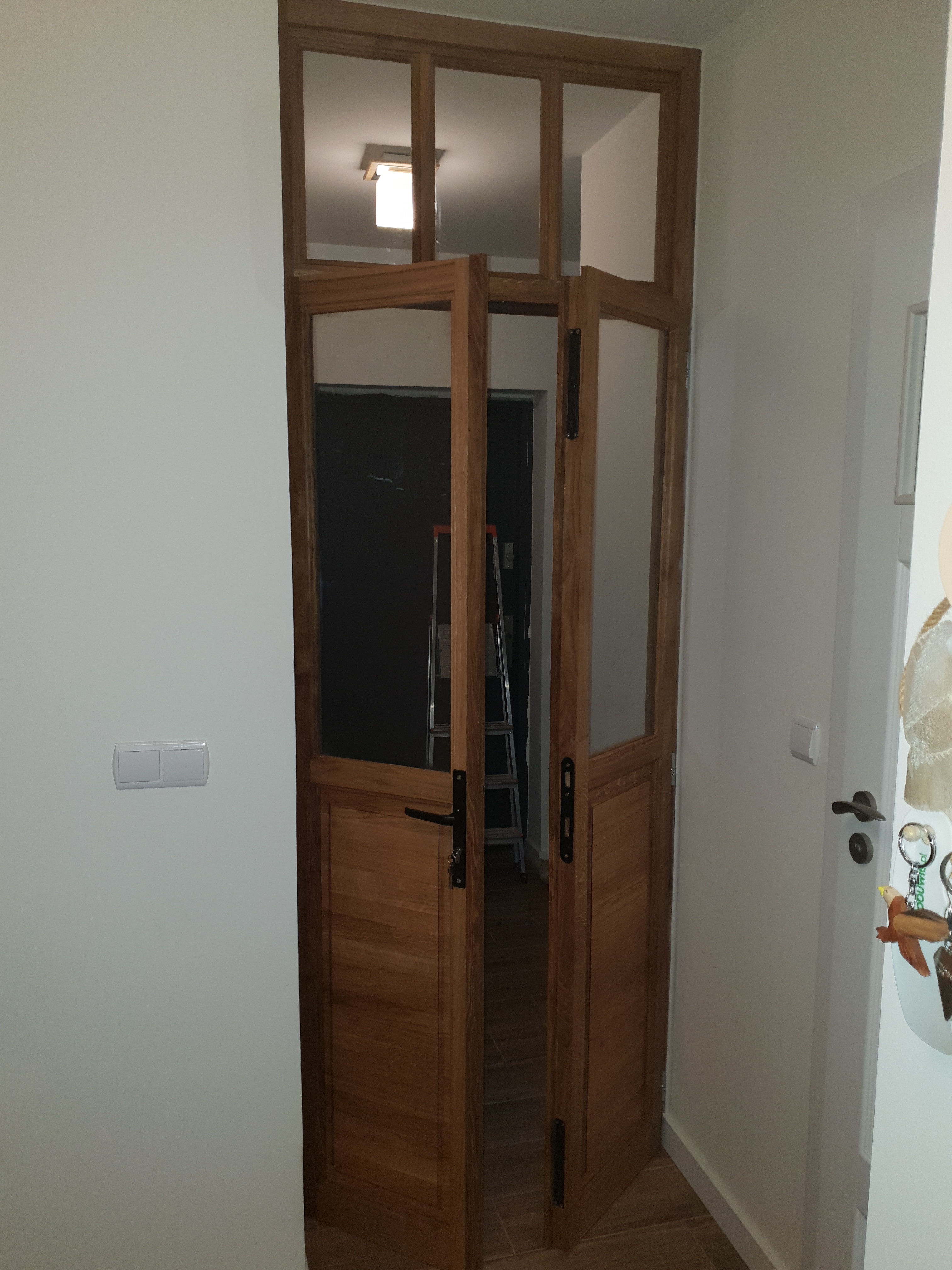 Otwarta, drewniana dwuskrzydłowa brama z przeszkleniami, zamontowana w jasnym wnętrzu w Krakowie, z widoczną drabiną w tle.