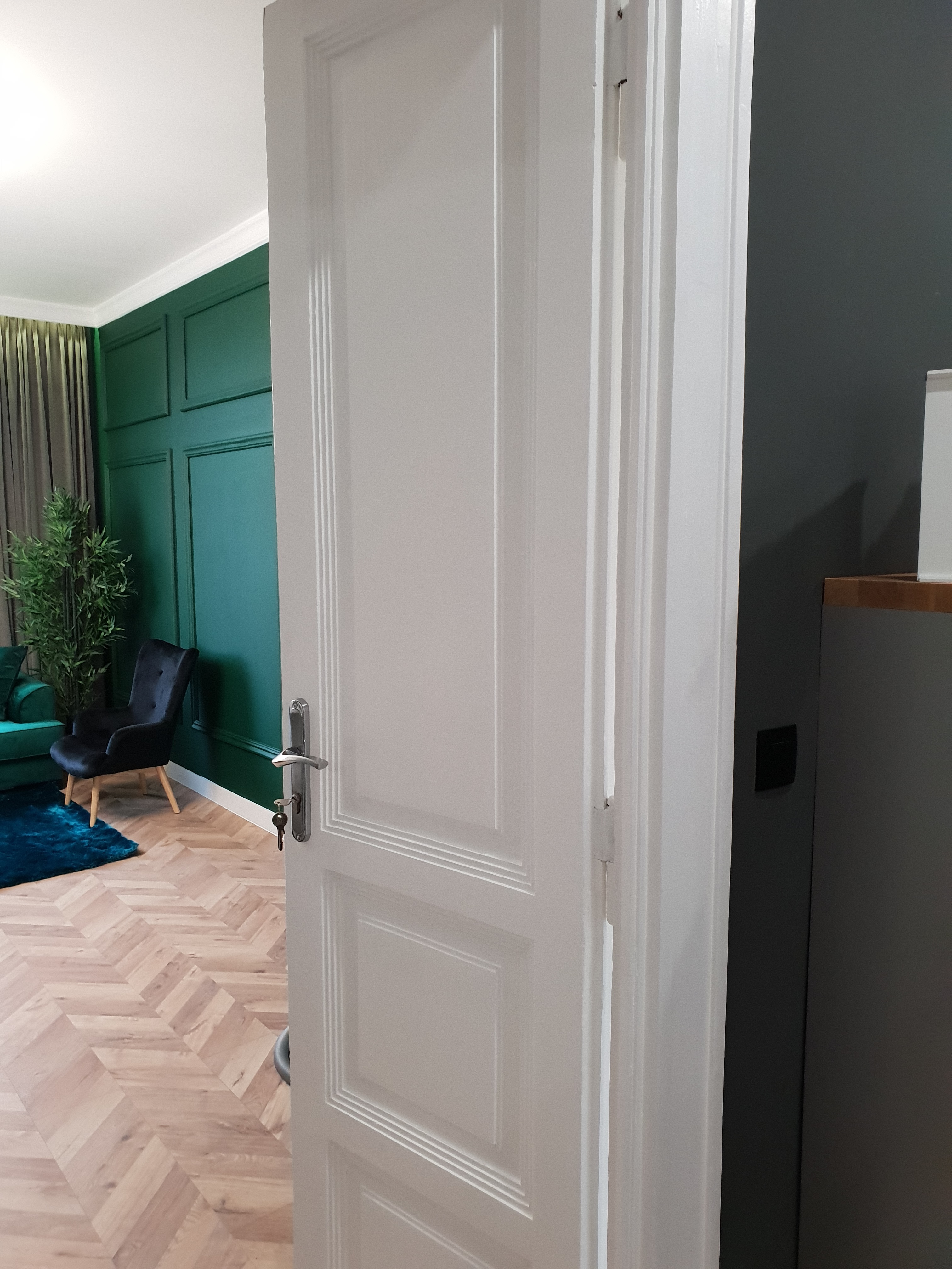 Otwarta biała, płycinowa konstrukcja z metalową klamką i kluczem w zamku na tle pokoju z zielonymi ścianami, parkietem w jodełkę i ciemnym fotelem.