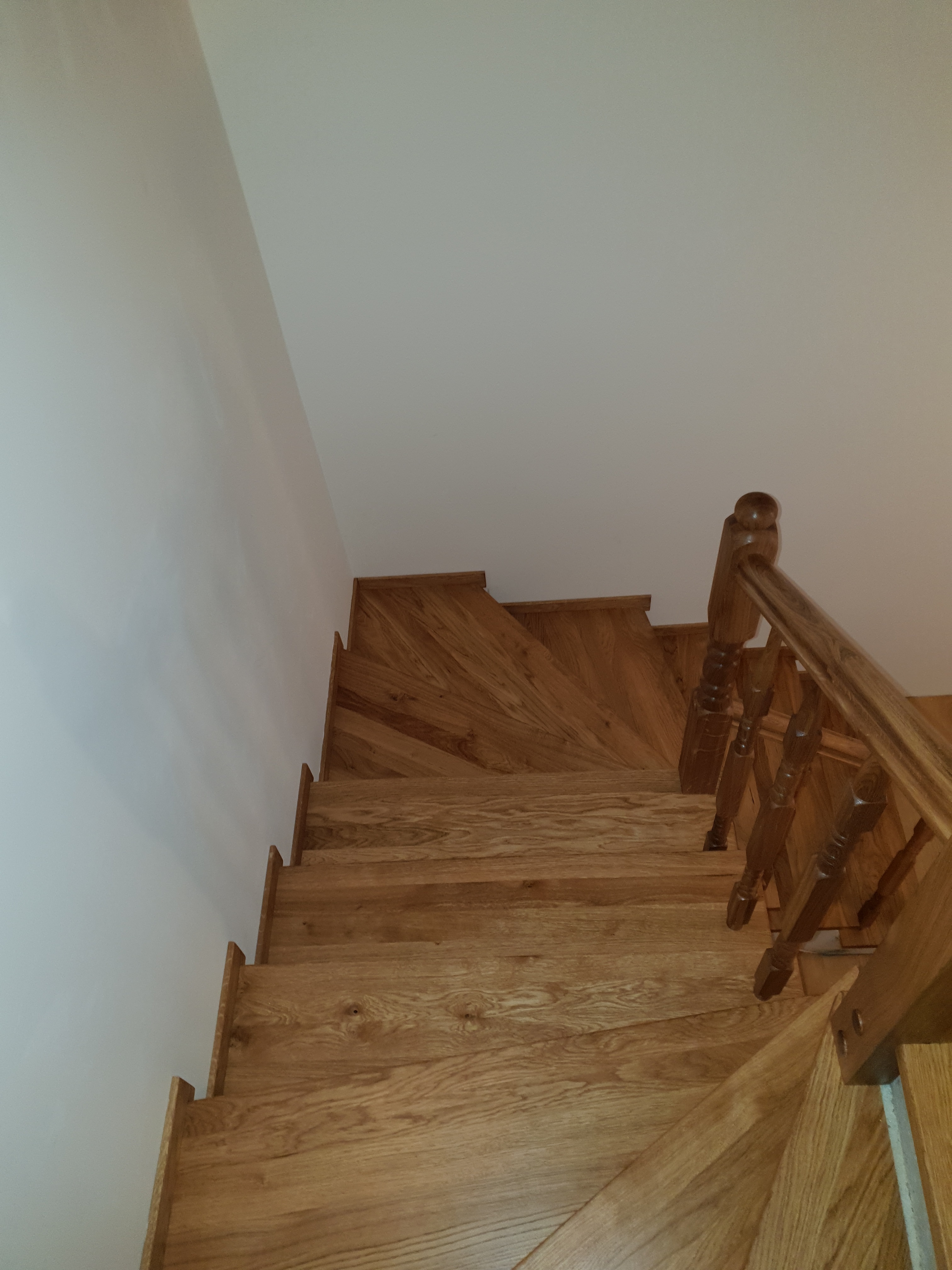 Kątowe ujęcie drewnianych schodów z balustradą, widok z góry na stopnie o naturalnym odcieniu drewna, przy białej ścianie.