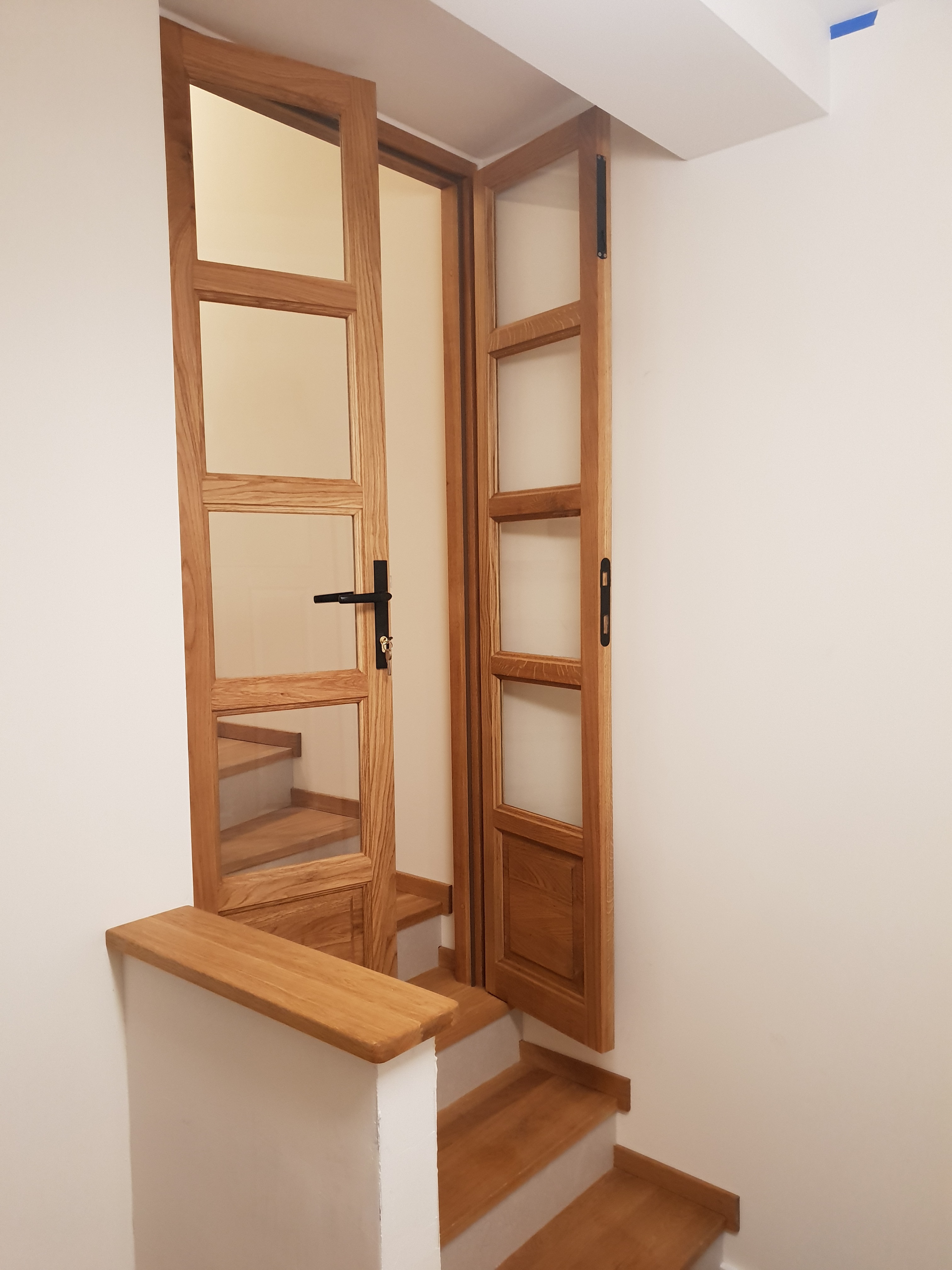 Drewniane, przeszklone drzwi wewnętrzne zamontowane przy schodach, z czarnymi klamkami, częściowo otwarte, ukazujące kolejne stopnie schodów.