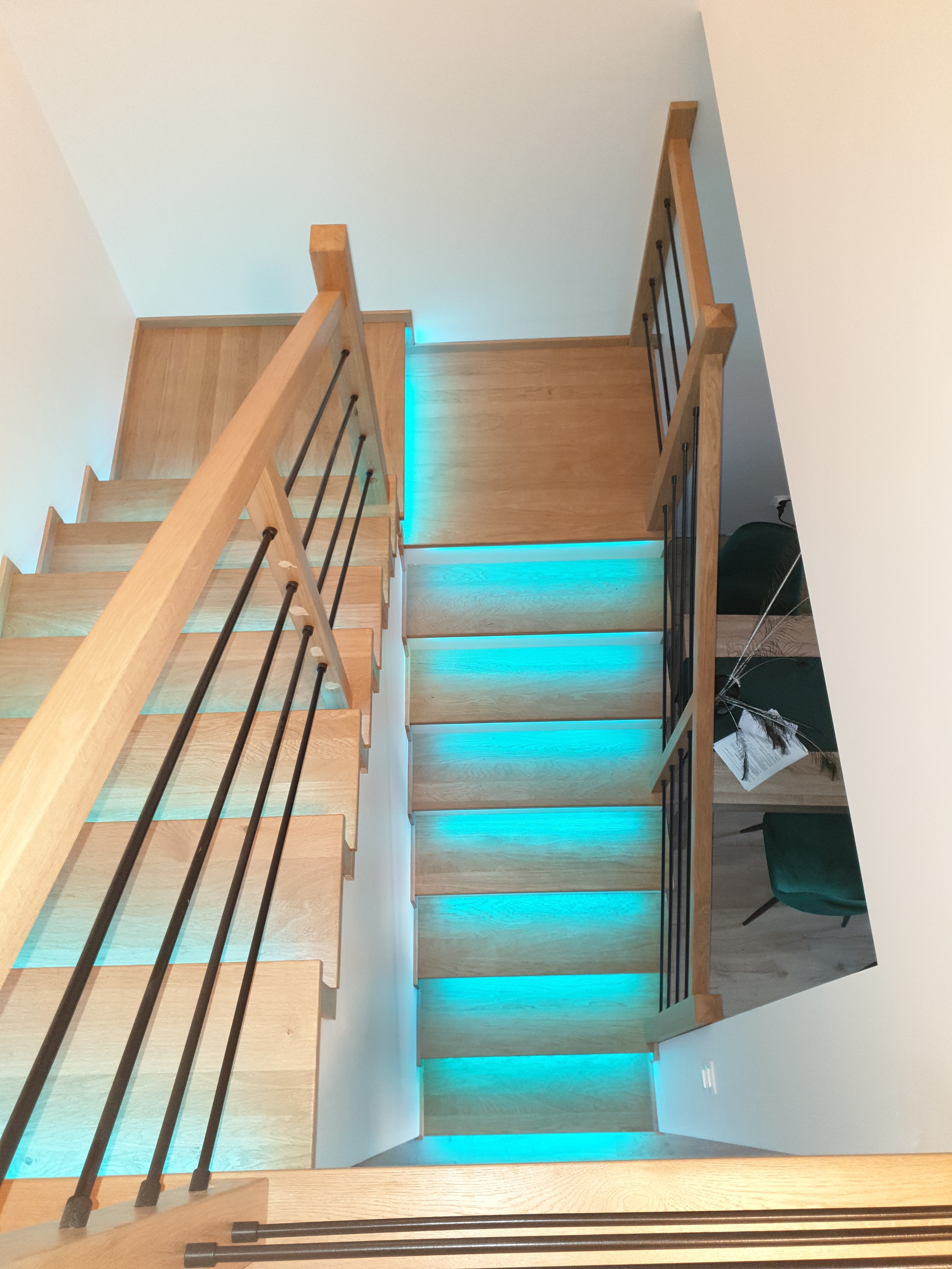 Nowoczesne schody drewniane z podświetleniem LED w kolorze turkusowym, widok z góry, czarne metalowe balustrady, jasne drewno, minimalistyczny design.