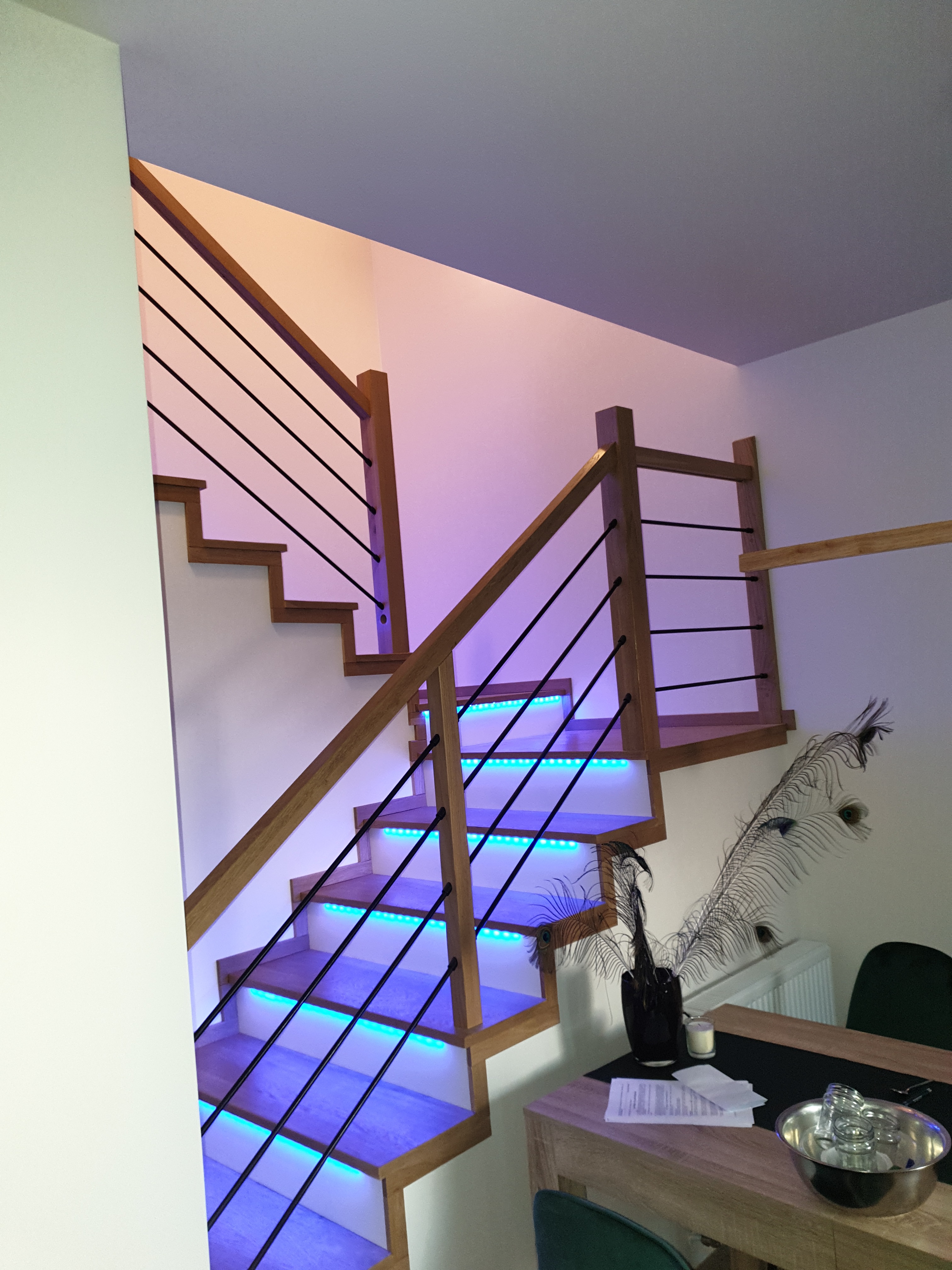Drewniane schody z czarną balustradą i podświetleniem LED w kolorze niebieskim, widok z dołu, dekoracyjne pióra pawie na stole w tle.