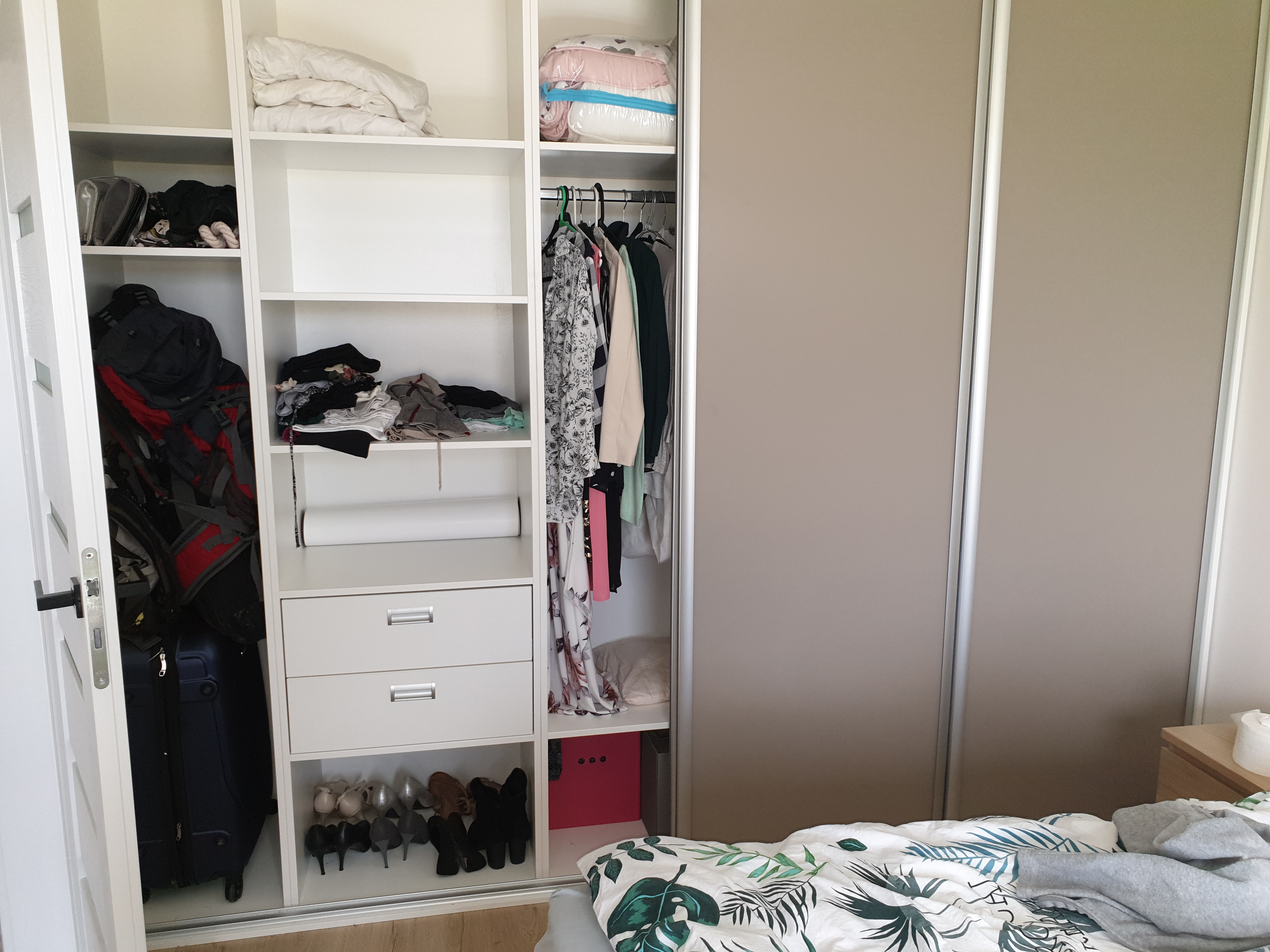 Wnętrze szafy wnękowej z przesuwnymi drzwiami w odcieniach beżu, widoczne półki z ubraniami, walizką, plecakiem i butami, białe szuflady i pudełko.