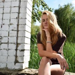 PhotoOK - Portret młodej kobiety z blond włosami, siedzącej na kamieniu obok białej, ceglanej ściany, z zielonymi roślinami w tle.