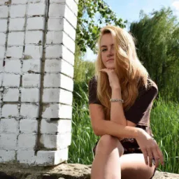 Portret młodej kobiety z blond włosami, siedzącej na kamieniu obok białej, ceglanej ściany, z zielonymi roślinami w tle.