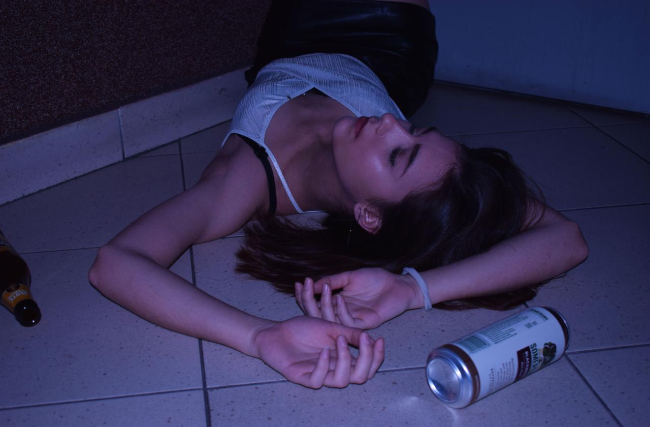 Młoda kobieta leżąca na podłodze w blasku neonu, obok puszki i butelki, z zamkniętymi oczami i dłońmi ułożonymi obok siebie.