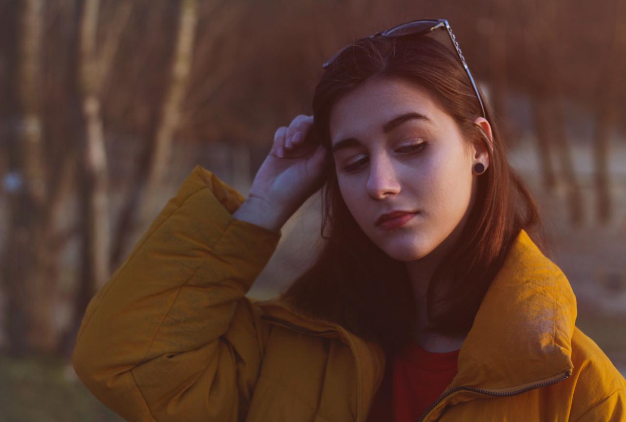 Portret młodej kobiety zamyślonej, w żółtej kurtce na tle drzew, okulary na głowie, delikatne światło.