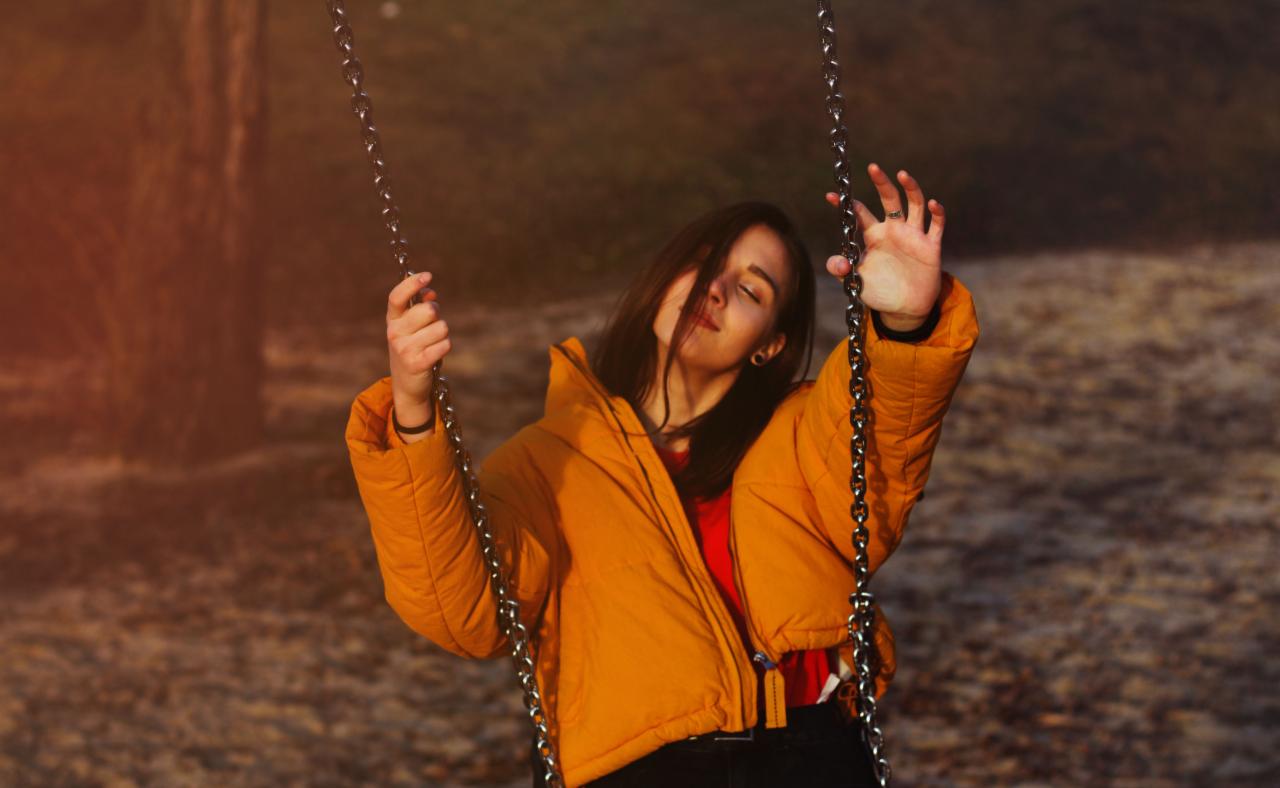 Młoda kobieta w pomarańczowej kurtce na huśtawce, z przymkniętymi oczami i wyciągniętą dłonią, uchwycona w jesiennej scenerii.