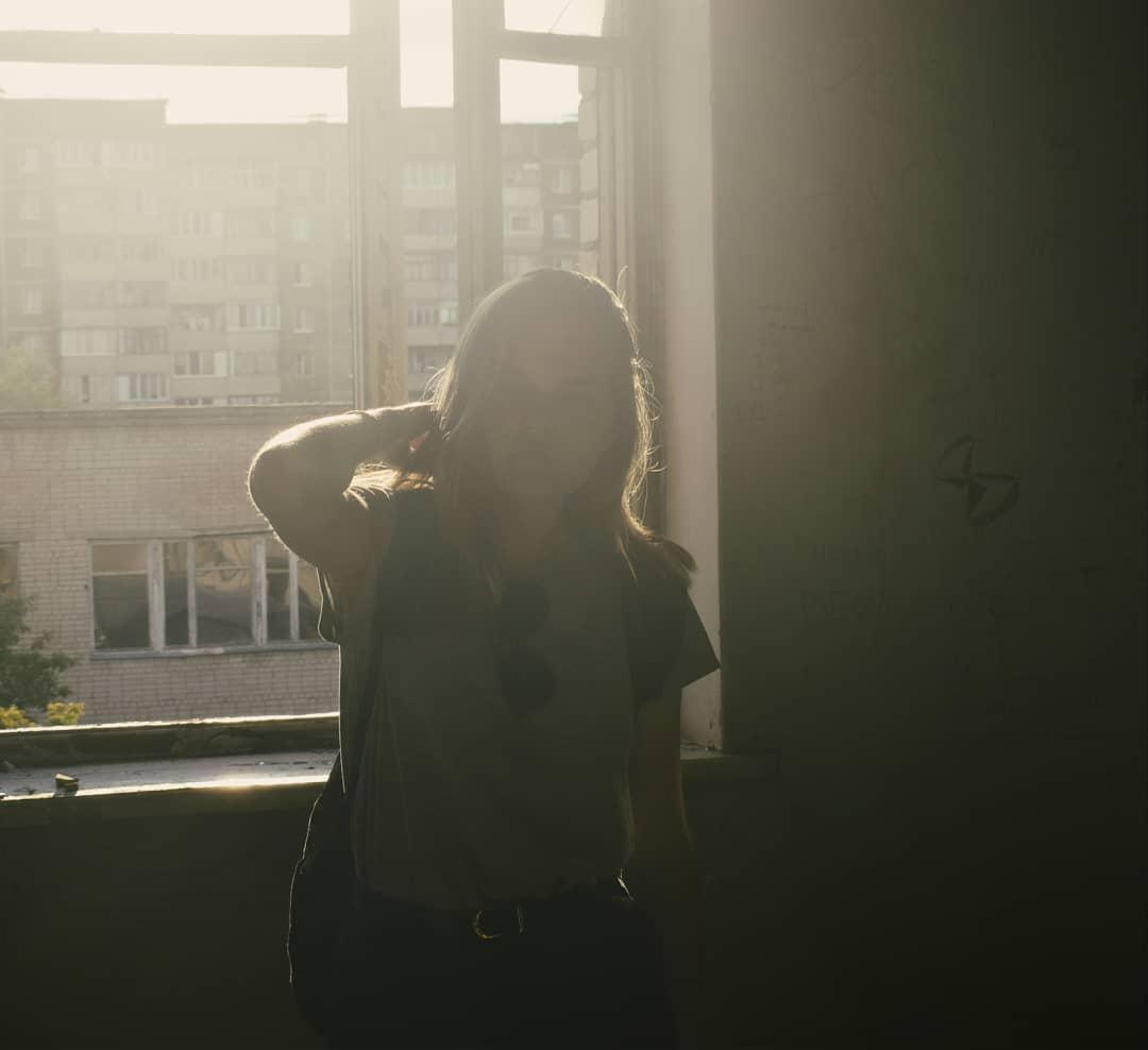 Kobieta stojąca w zrujnowanym wnętrzu, oświetlona mocnym światłem wpadającym przez okno, na ścianie widoczne graffiti.