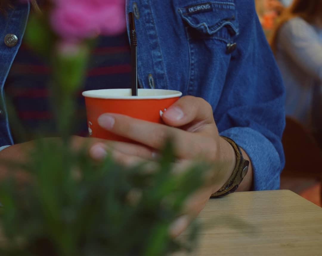 Osoba trzymająca pomarańczowy kubek z napojem i słomką, ubrana w dżinsową kurtkę, z rozmytym kwiatem i rośliną w tle.