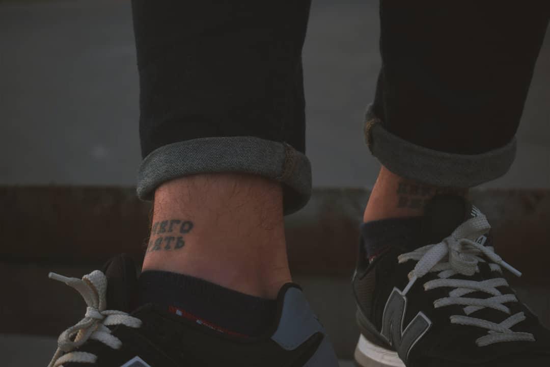 Nogi w dżinsach i sportowych butach, tatuaże na kostkach z rosyjskimi napisami.
