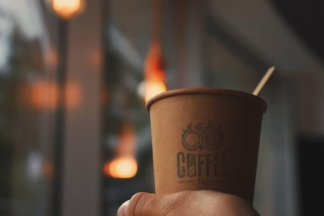 Papierowy kubek z kawą, z logo i drewnianym mieszadełkiem, trzymany w dłoni na rozmytym tle z żarówkami.