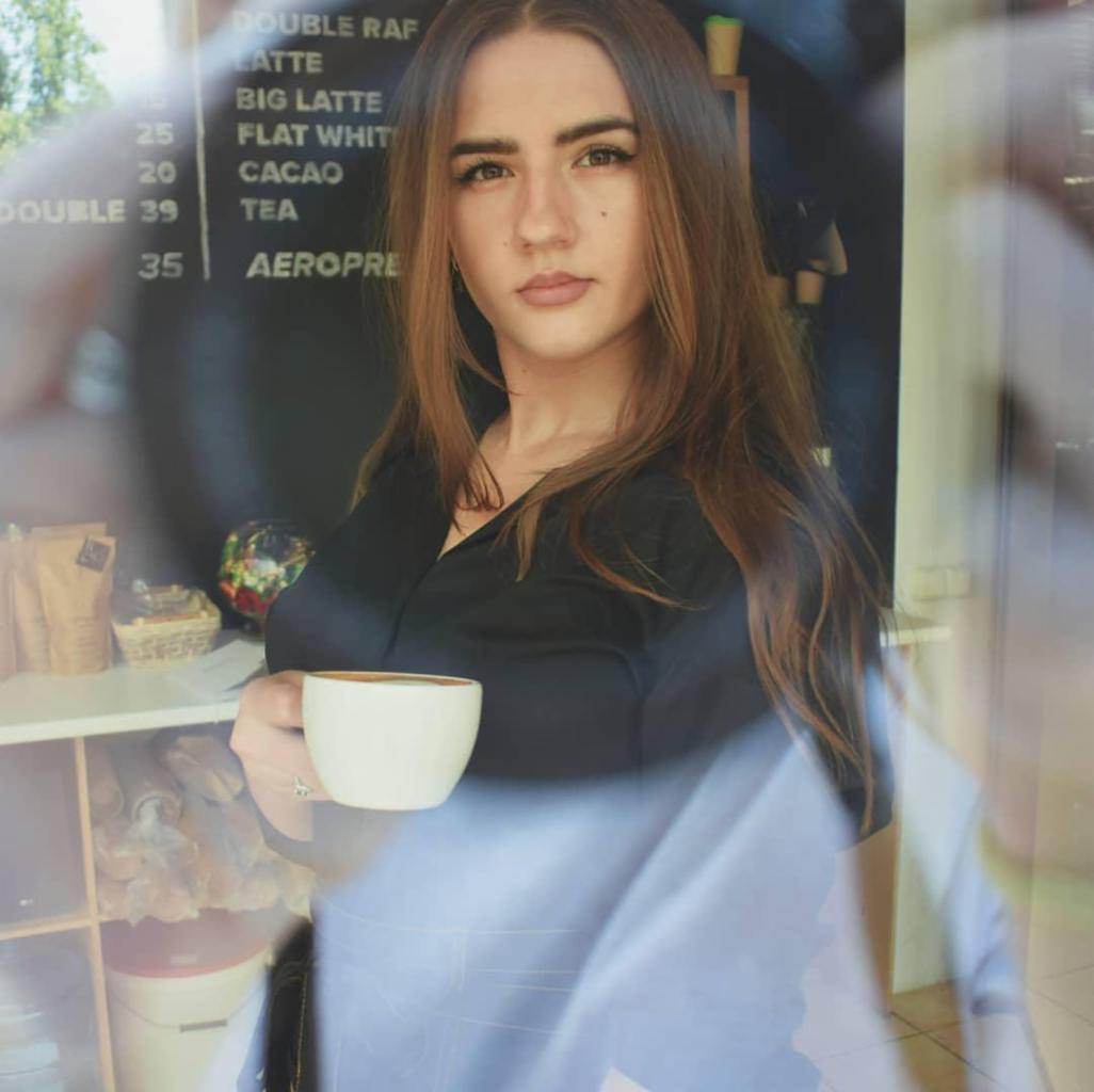 Młoda kobieta w Opolu, ubrana w czarną koszulę, trzyma białą filiżankę kawy na tle tablicy z menu kawiarni.