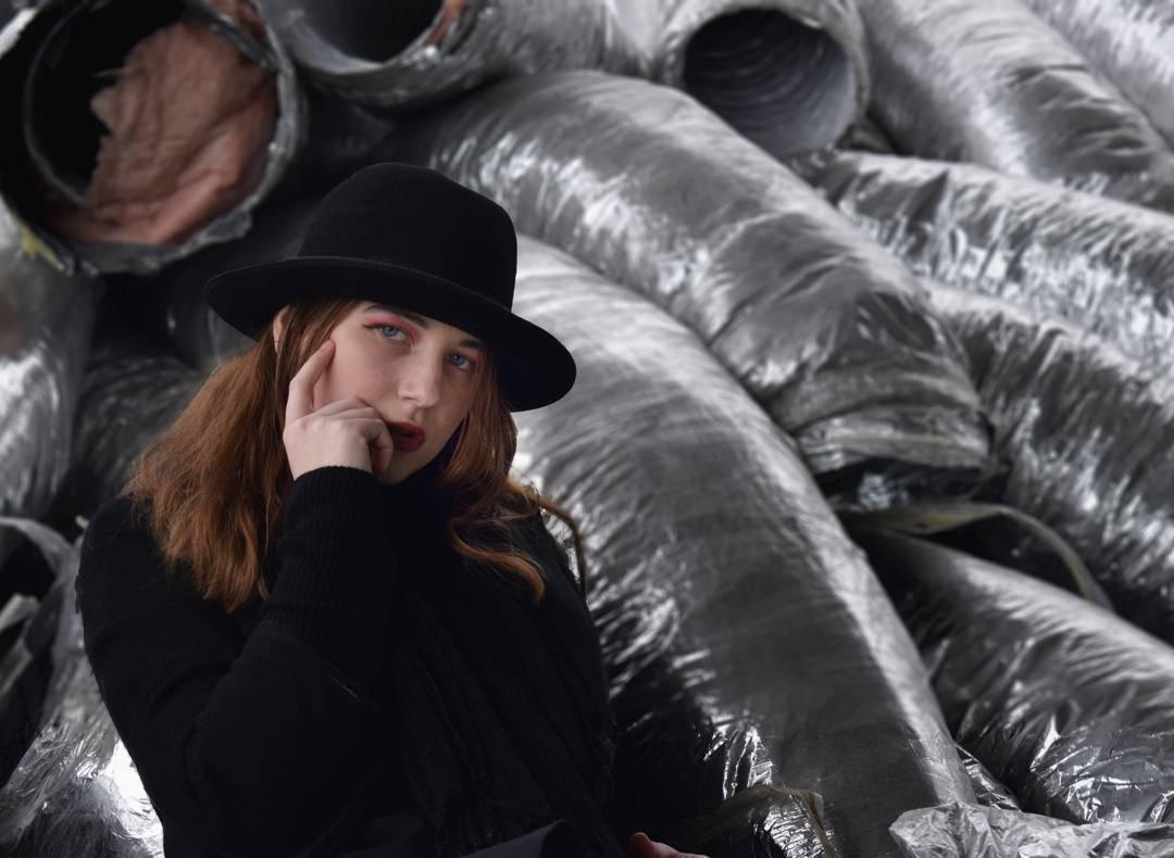 Portret kobiety w czarnym kapeluszu na tle stosu srebrnych, izolowanych rur wentylacyjnych