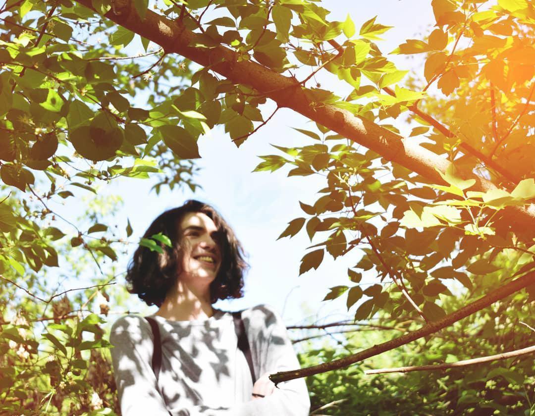 Uśmiechnięta młoda kobieta z ciemnymi włosami, sfotografowana z dołu przez gałęzie drzew w słoneczny dzień, z liściem we włosach.