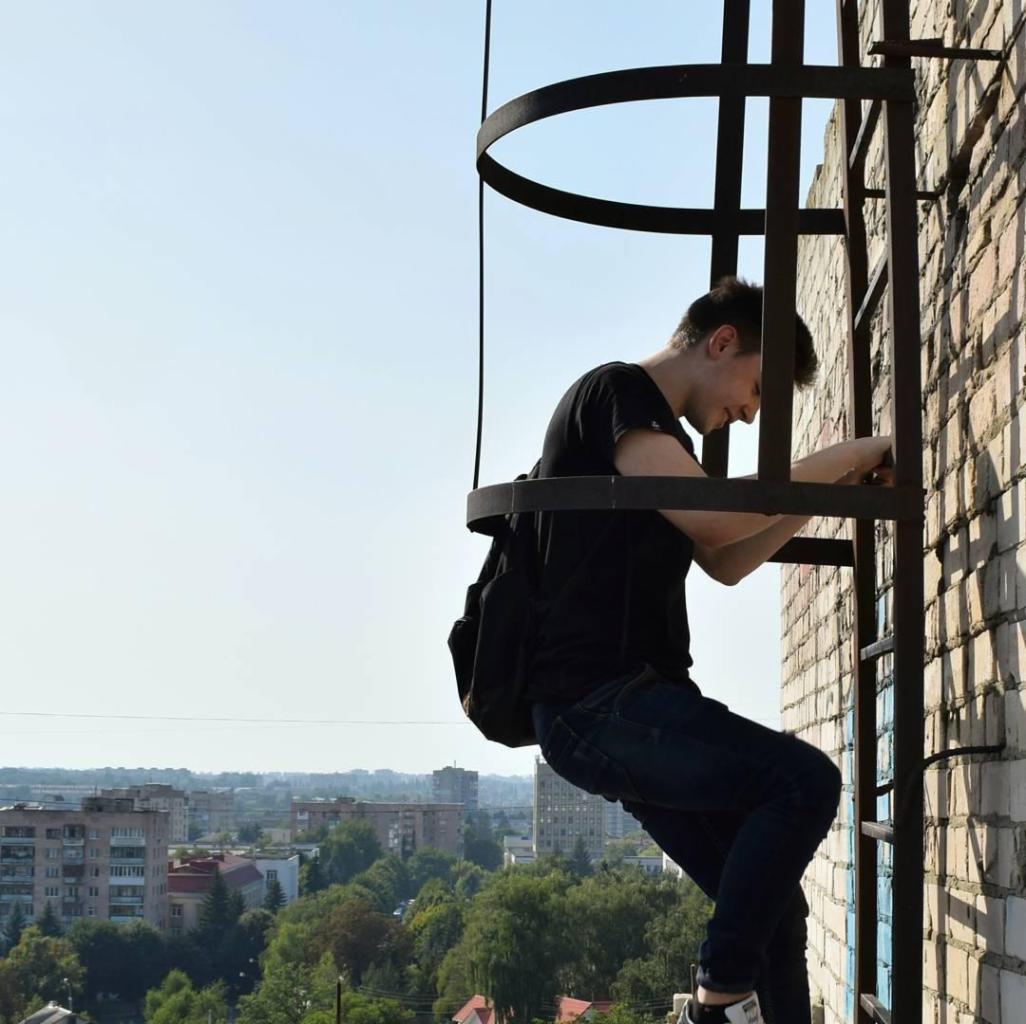Młody mężczyzna w czarnej koszulce i jeansach wspina się po metalowej drabinie przymocowanej do ceglanego budynku, z widokiem na miasto w tle.