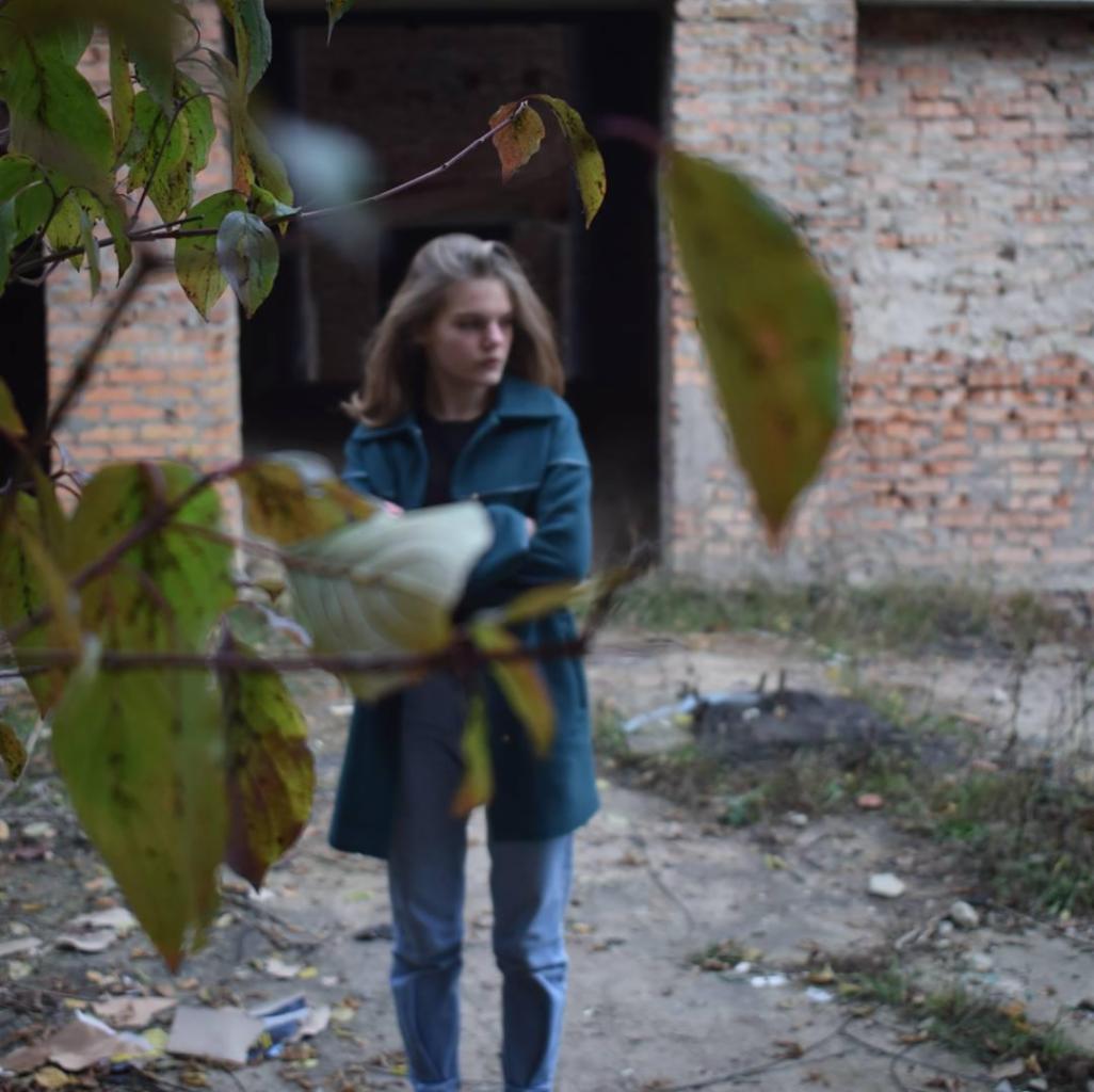 Młoda kobieta w zielonym płaszczu pozuje na tle ceglanego budynku w trakcie sesji kobiecej, z liśćmi na pierwszym planie.