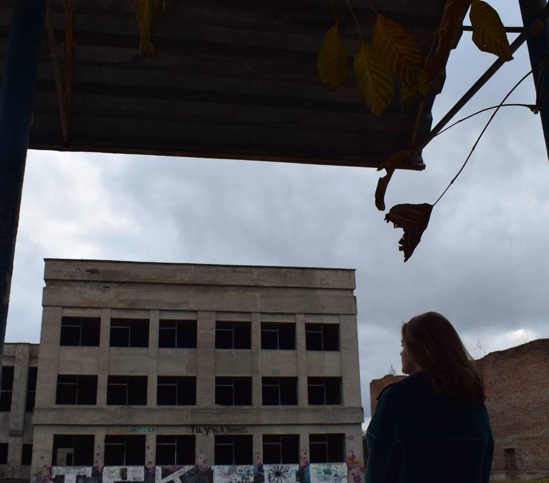 Kobieta spogląda na opuszczony budynek z graffiti, widziana spod zadaszenia z wiszącymi suchymi liśćmi.