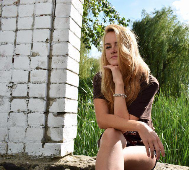 Portret młodej kobiety z blond włosami, siedzącej na kamieniu obok białej, ceglanej ściany, z zielonymi roślinami w tle.