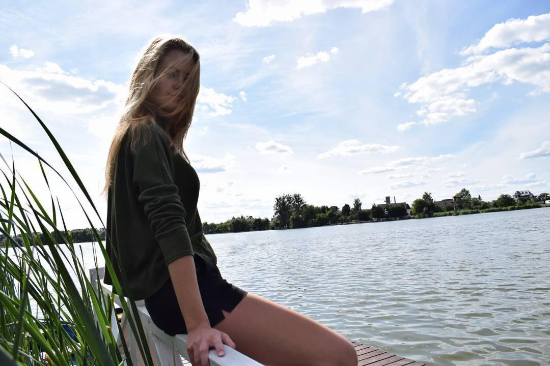 Młoda kobieta siedzi na drewnianym pomoście nad jeziorem, wiatr rozwiewa jej włosy, w tle błękitne niebo z chmurami i zieleń drzew.