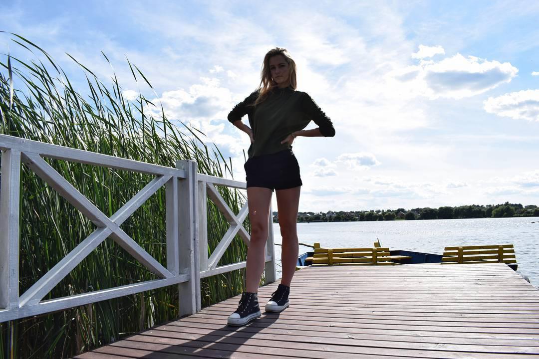 Młoda kobieta w ciemnozielonej bluzce i czarnych szortach pozuje na drewnianym molo z rękami na biodrach, w tle jezioro, trzciny i błękitne niebo z chmurami.