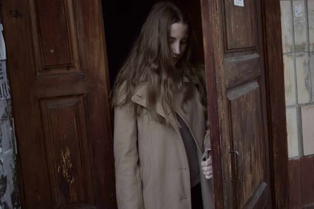 Kobieta w beżowym płaszczu wychodzi przez drewniane drzwi z widocznymi śladami użytkowania, trzymając za klamkę.