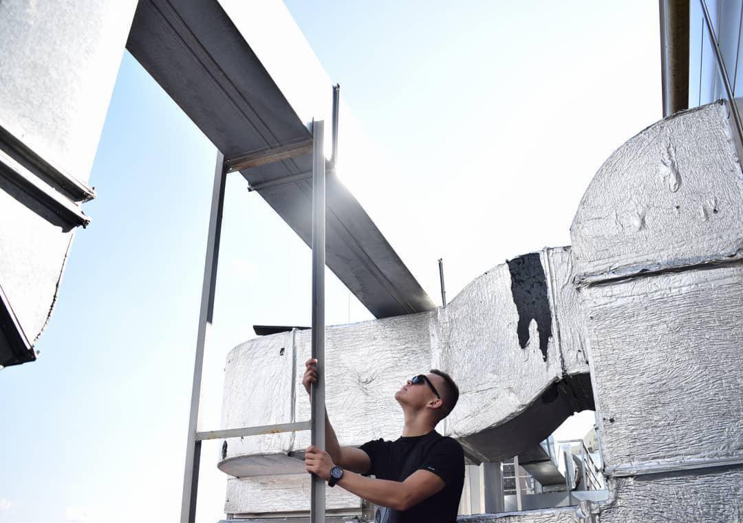 Mężczyzna w okularach przeciwsłonecznych wspina się po metalowej konstrukcji, prawdopodobnie podczas instalacji lub konserwacji systemu wentylacyjnego, widoczne izolowane kanały wentylacyjne...