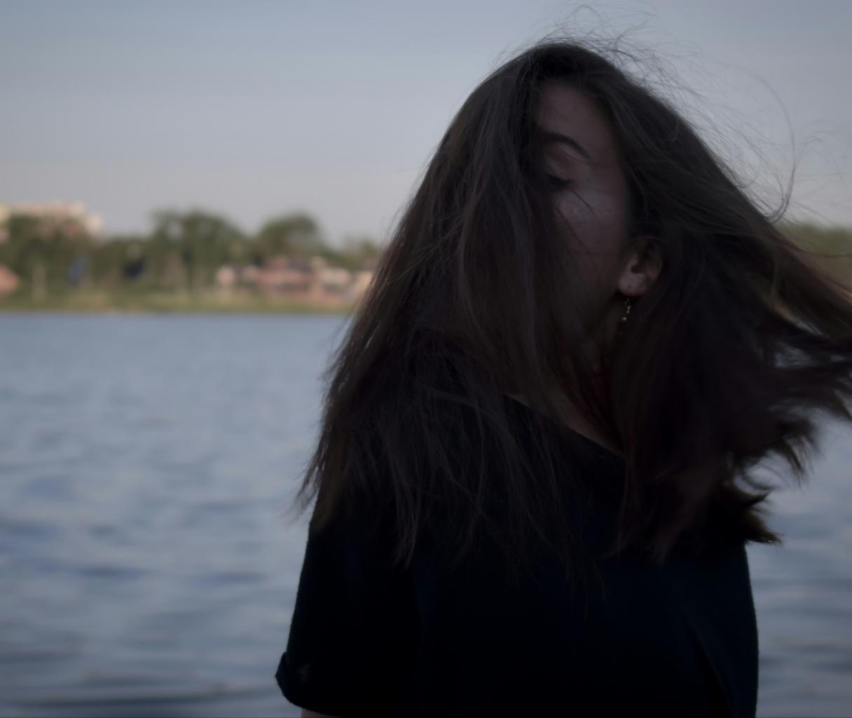 Portret kobiety z długimi, ciemnymi włosami zasłaniającymi twarz, na tle jeziora i oddalonych budynków w spokojnej, stonowanej kolorystyce.