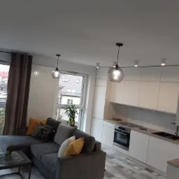 Nowoczesny salon z aneksem kuchennym: szara sofa narożna z dekoracyjnymi poduszkami, stolik kawowy, biała zabudowa kuchenna z czarnym zlewem i płytą indukcyjną, okno z widokiem na zewnątrz, lampy...