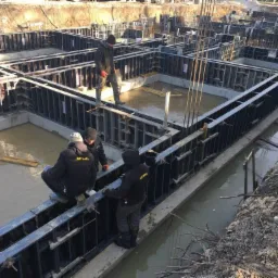 Budowa fundamentów domu: pracownicy w ciemnych ubraniach na metalowej konstrukcji szalunkowej, w tle zalane wodą wykopy pod fundamenty, widoczne stalowe pręty zbrojeniowe.