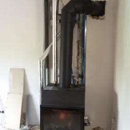 Częściowo zamontowany czarny piec kominkowy z widocznymi przewodami spalinowymi, instalacją wodną i elementami konstrukcyjnymi w surowym wnętrzu.