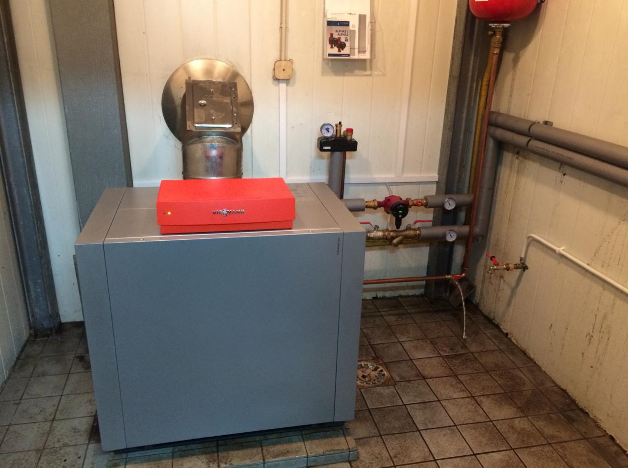 Szary piec grzewczy Viessmann z czerwonym panelem sterowania w kotłowni z ceramiczną podłogą, widoczne rury i zawory instalacji grzewczej oraz komin spalinowy.