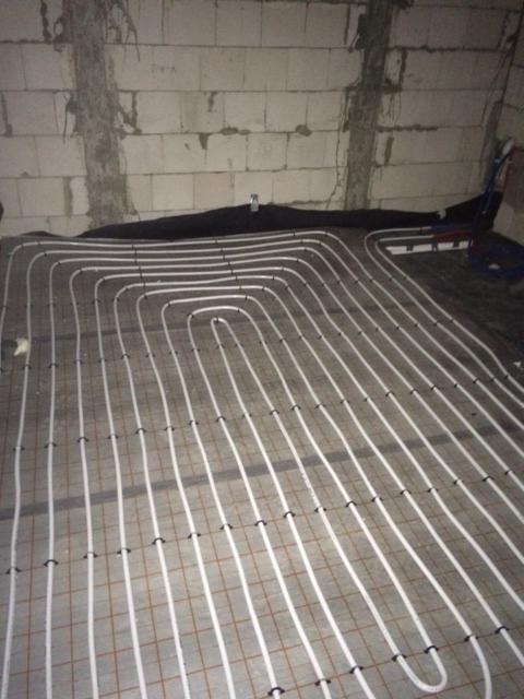 Białe rury układane w pętlach na siatce podłogowej, przygotowane do zalania betonem, surowa ściana z bloczków w tle.