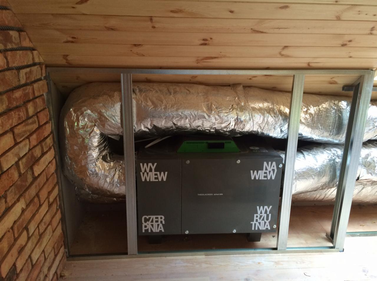Szara jednostka rekuperacyjna Thessla Green AirPack 500N z widocznymi przewodami wentylacyjnymi w izolacji aluminiowej, umieszczona w przestrzeni poddasza z drewnianym wykończeniem i fragmentem...