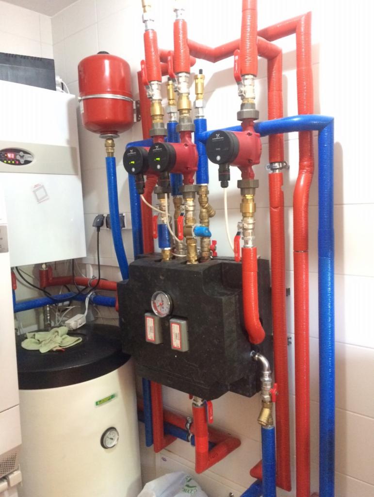 Złożony system rur grzewczych w kolorach czerwonym i niebieskim, z trzema pompami obiegowymi Wilo, zbiornikiem wyrównawczym i kotłem grzewczym na tle białych płytek.