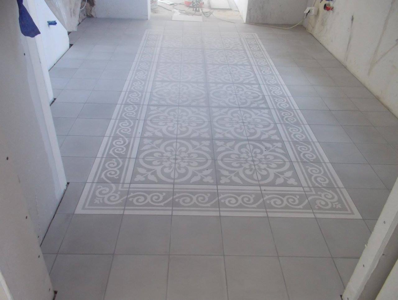 Szare płytki podłogowe z dekoracyjnym wzorem w stylu vintage, ułożone w prostokąt na środku pomieszczenia w trakcie remontu.