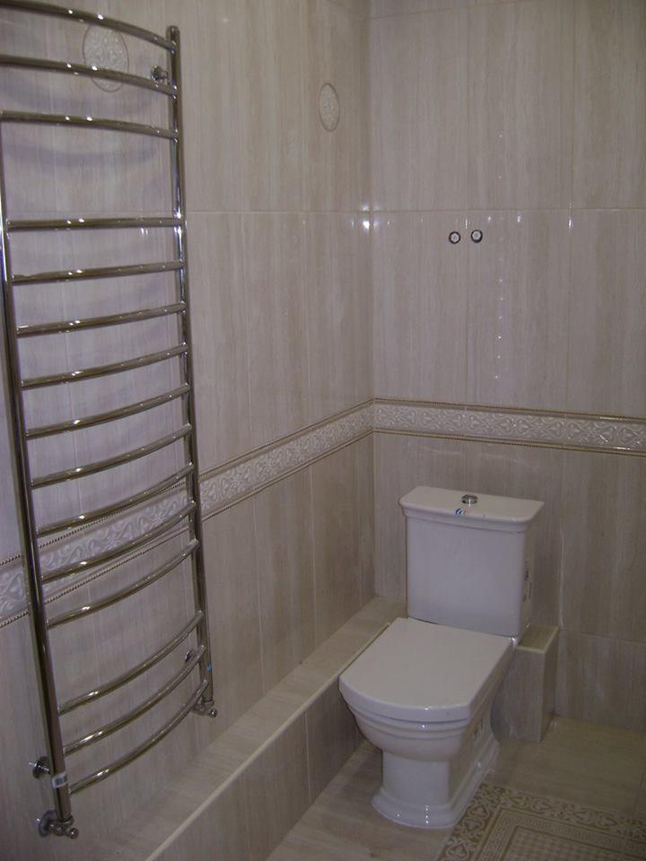 Wykończona łazienka z beżowymi płytkami, grzejnikiem drabinkowym i toaletą z ceramiczną spłuczką.