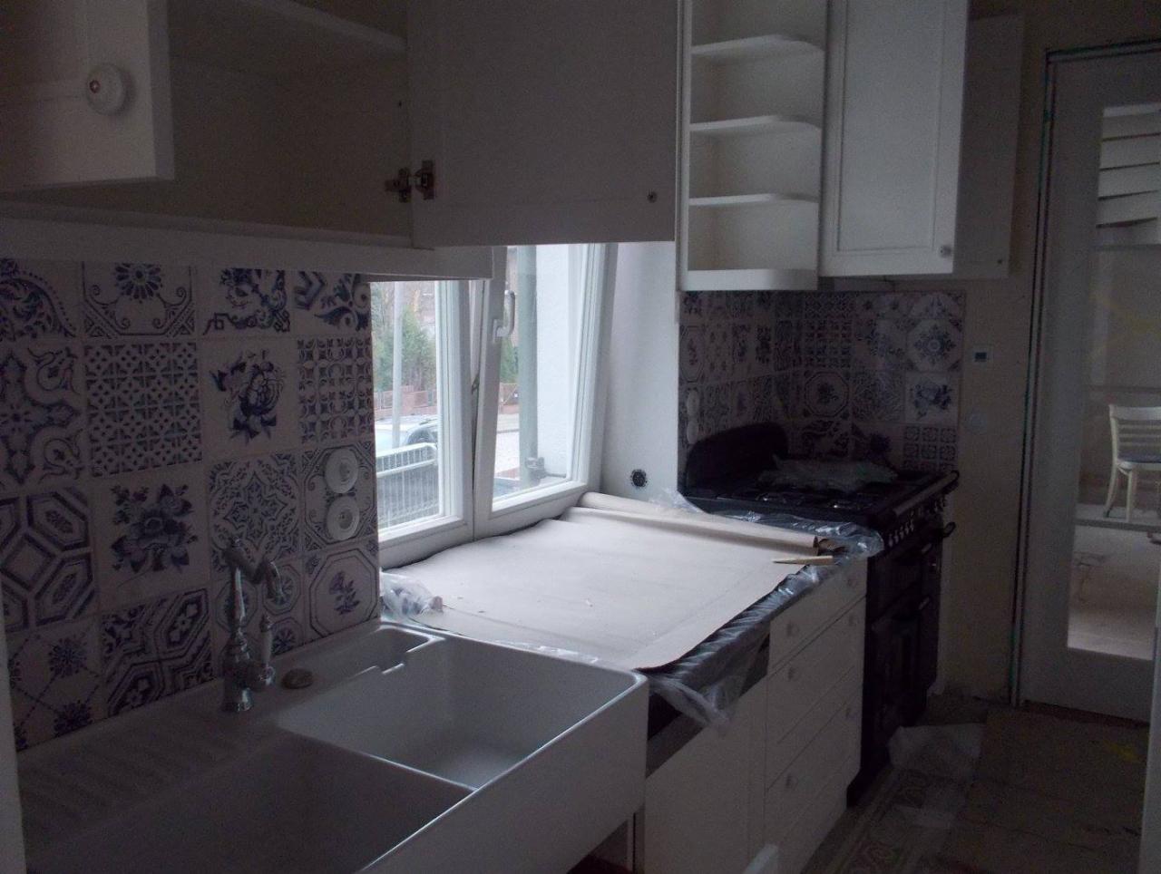 Kuchnia w trakcie remontu: białe szafki wiszące, ceramiczna umywalka z ozdobną baterią, ściana wyłożona dekoracyjnymi płytkami w odcieniach bieli i niebieskiego, blat zabezpieczony folią ochronną.